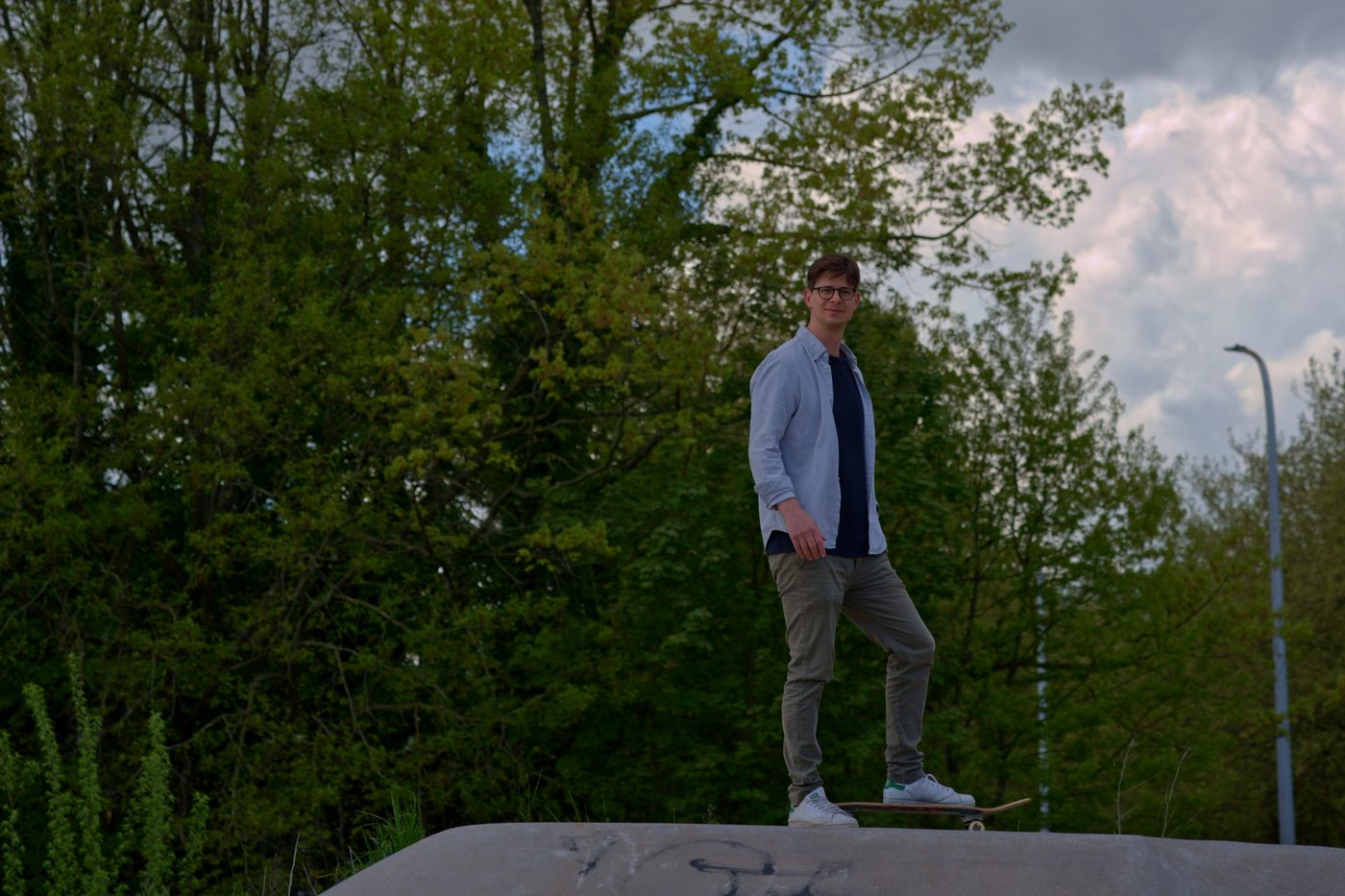 Portrait en extérieur d'un client debout sur son skateboard dans un skatepark, arbres et ciel d'orage en arrière-plan